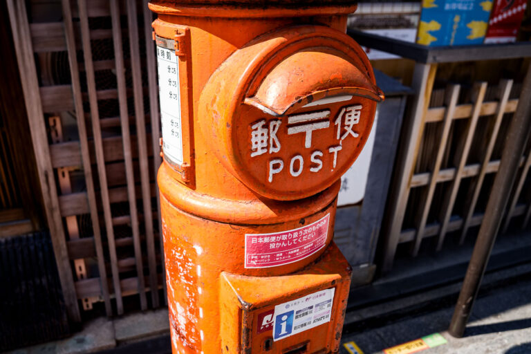 Vintage Japanese Post Box in Kyoto 4 A cylindrical orange post box on a street in Kyoto, photographed in March 2023. This style of mailbox—introduced in the early 1900s during Japan’s push to modernize its postal infrastructure—became a familiar feature across the country through the mid-20th century. Its rounded form and front mail slot were designed to be durable, weather-resistant, and easy to recognize even in densely built neighborhoods.Although newer rectangular mailboxes are now common in urban areas, many historic districts in Kyoto retain these older designs to preserve the traditional streetscape. Today, they continue to function as part of Japan Post’s network, blending everyday utility with a tangible link to the city’s architectural and civic history.