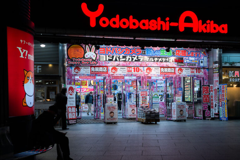 Yodobashi-Akiba Exterior at Night 3 Yodobashi-Akiba, photographed in March 2023, is one of Tokyo’s largest electronics retail complexes and a major landmark in the Akihabara district. Opened in 2005 as part of a wider redevelopment effort around JR Akihabara Station, the multi-story store consolidated several specialty floors—computers, cameras, gaming, home appliances, musical instruments—into a single destination that helped redefine the area’s retail identity in the 2000s.Its brightly illuminated entrance, typically covered in seasonal and promotional signage, reflects the competitive electronics market that shaped modern Akihabara. The building also connects directly to the Tsukuba Express line and sits beside the redevelopment zone that transformed the district from its postwar reputation as “Electric Town” into a mix of technology retail, office towers, and entertainment venues.