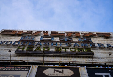 The iconic neon sign of Yodobashi Camera’s head store in Shinjuku is seen from street level, with its mix of Japanese and English lettering. The large retro-style signage, lined with anti-bird spikes, reflects the store’s long-standing presence as one of Tokyo’s biggest electronics retailers. Yodobashi Camera’s Shinjuku West Exit location serves as the flagship branch, anchoring the city’s vast camera and tech shopping district.