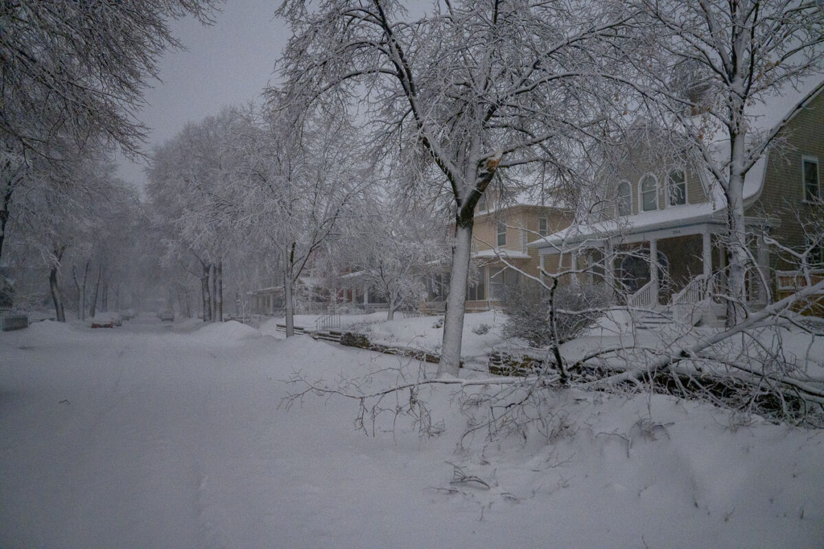 A snow-covered residential street in Minneapolis during an April 2023 blizzard, with homes and snow-laden trees lining the road.