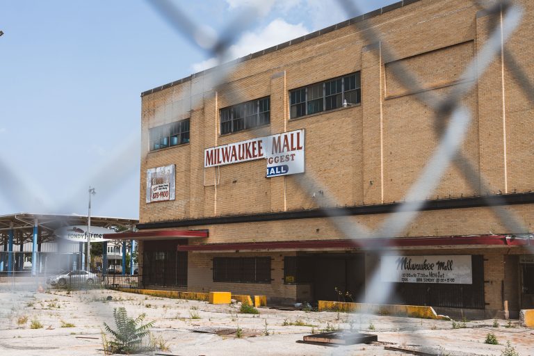 Milwaukee Mall Sits Vacant Behind a Chain Link Fence 4 The abandoned Milwaukee Mall building in Wisconsin stands vacant with boarded windows and overgrown pavement.
