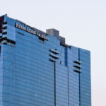 Hilton Grand Vacations at Resorts World Las Vegas is a sleek, glass-covered tower on the northern end of the Strip, known for its reflective façade and distinctive red architectural accent running along one edge. Completed in 2021 as part of the Resorts World complex, the property offers timeshare-style accommodations within one of the newest integrated resorts in the city. Its elevated design provides sweeping views of the Strip and nearby mountains, while its proximity to the Las Vegas Convention Center and the airport—seen in the distance here—highlights its mix of leisure and business appeal.