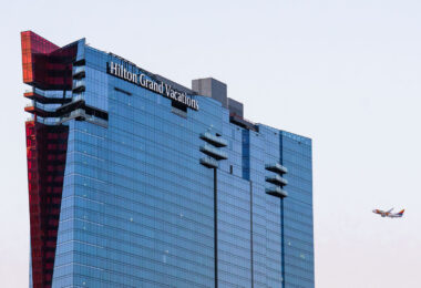 Hilton Grand Vacations at Resorts World Las Vegas is a sleek, glass-covered tower on the northern end of the Strip, known for its reflective façade and distinctive red architectural accent running along one edge. Completed in 2021 as part of the Resorts World complex, the property offers timeshare-style accommodations within one of the newest integrated resorts in the city. Its elevated design provides sweeping views of the Strip and nearby mountains, while its proximity to the Las Vegas Convention Center and the airport—seen in the distance here—highlights its mix of leisure and business appeal.