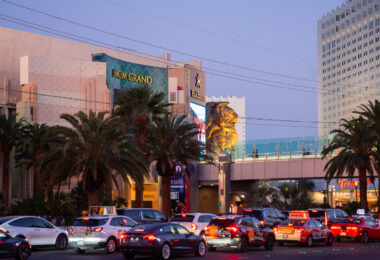 The MGM Grand stands as one of the most iconic landmarks on the Las Vegas Strip, instantly recognizable by its emerald-green lighting and massive golden lion statue at the entrance. Opened in 1993, it remains one of the largest hotels in the world, housing over 6,800 rooms, a sprawling casino, and major entertainment venues. The resort is home to Hakkasan nightclub and has hosted countless concerts, boxing matches, and celebrity residencies over the years. In this scene, evening traffic and palm trees frame the grand façade, capturing the constant motion and energy that define this busy corner of Las Vegas Boulevard.