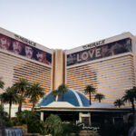 The Mirage and The Beatles LOVE, Las Vegas 1 The exterior of The Mirage on the Las Vegas Strip, photographed with the resort’s twin hotel towers and central entrance framed by palm landscaping and the property’s signature tropical aesthetic. Prominently displayed along the upper façades are large-format banners for The Beatles LOVE, the long-running Cirque du Soleil production that opened in 2006 and became one of the Strip’s most recognizable resident shows.Opened in 1989 by Steve Wynn, The Mirage marked a turning point in modern Las Vegas resort development, helping shift the Strip toward large-scale, theme-driven destinations that combined lodging, entertainment, and spectacle into a single integrated property. The resort’s gold-tinted glass towers, central dome, and lush frontage were designed to evoke a self-contained oasis amid the surrounding desert and high-density casino corridor. For nearly two decades, LOVE tied the hotel’s identity to the legacy of The Beatles, blending archival music with acrobatics, choreography, and immersive theater in a purpose-built venue inside the resort.