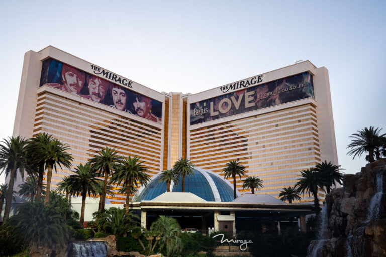 The Mirage and The Beatles LOVE, Las Vegas 1 The exterior of The Mirage on the Las Vegas Strip, photographed with the resort’s twin hotel towers and central entrance framed by palm landscaping and the property’s signature tropical aesthetic. Prominently displayed along the upper façades are large-format banners for The Beatles LOVE, the long-running Cirque du Soleil production that opened in 2006 and became one of the Strip’s most recognizable resident shows.Opened in 1989 by Steve Wynn, The Mirage marked a turning point in modern Las Vegas resort development, helping shift the Strip toward large-scale, theme-driven destinations that combined lodging, entertainment, and spectacle into a single integrated property. The resort’s gold-tinted glass towers, central dome, and lush frontage were designed to evoke a self-contained oasis amid the surrounding desert and high-density casino corridor. For nearly two decades, LOVE tied the hotel’s identity to the legacy of The Beatles, blending archival music with acrobatics, choreography, and immersive theater in a purpose-built venue inside the resort.