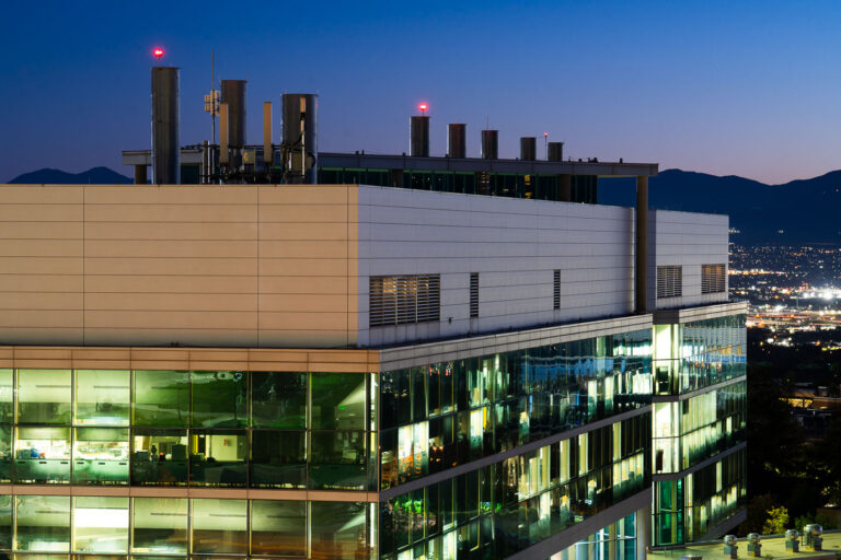 University of Utah Biomedical Engineering Building at Dusk 2 The University of Utah's Biomedical Engineering Building at dusk, featuring modern architecture with glass facades and illuminated interiors, set against a backdrop of distant city lights and mountains.
