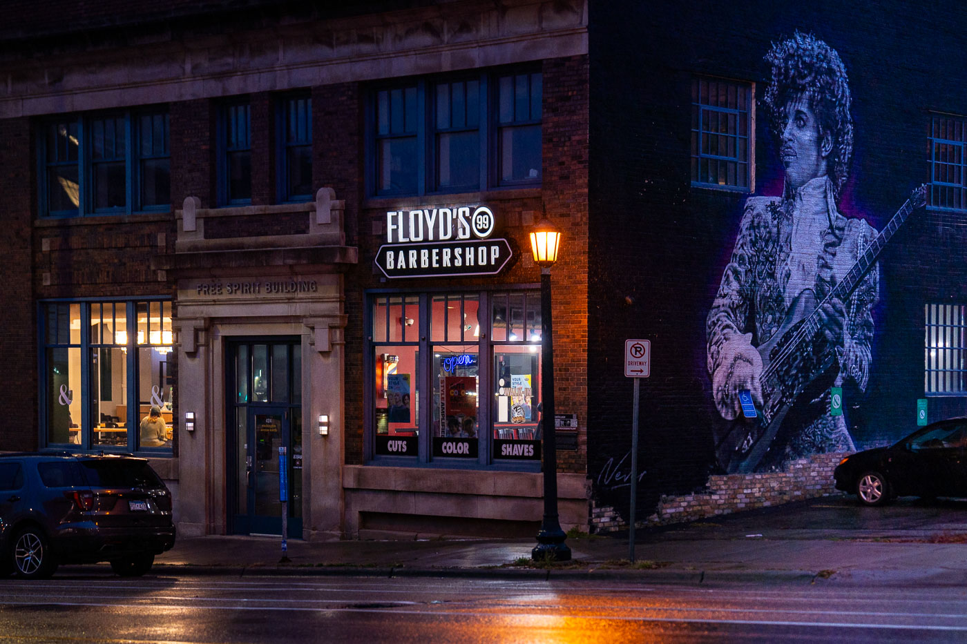 Floyd's 99 Barbershop on Washington Avenue North in Minneapolis's North Loop neighborhood, with a Prince mural on the adjacent brick wall, at night.