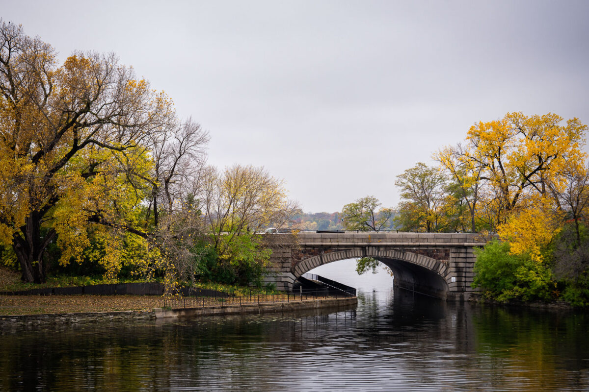 The Midtown Greenway Bridge in Minneapolis, a stone arch structure, carries traffic over a waterway surrounded by autumn foliage.