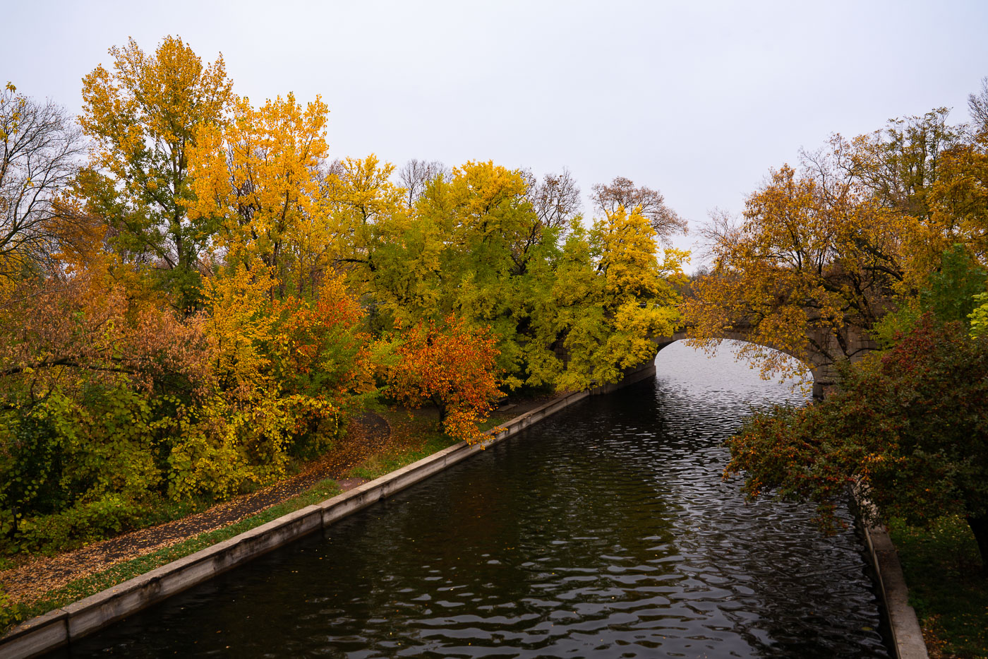 Midtown Greenway in Minneapolis, MN, features a canal with trees displaying autumn colors and an arched stone bridge in the background.