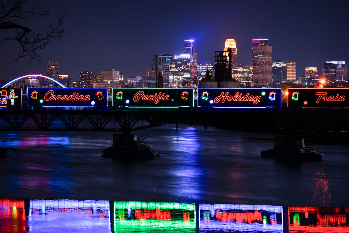 Lit up holiday train crosses river with skyline