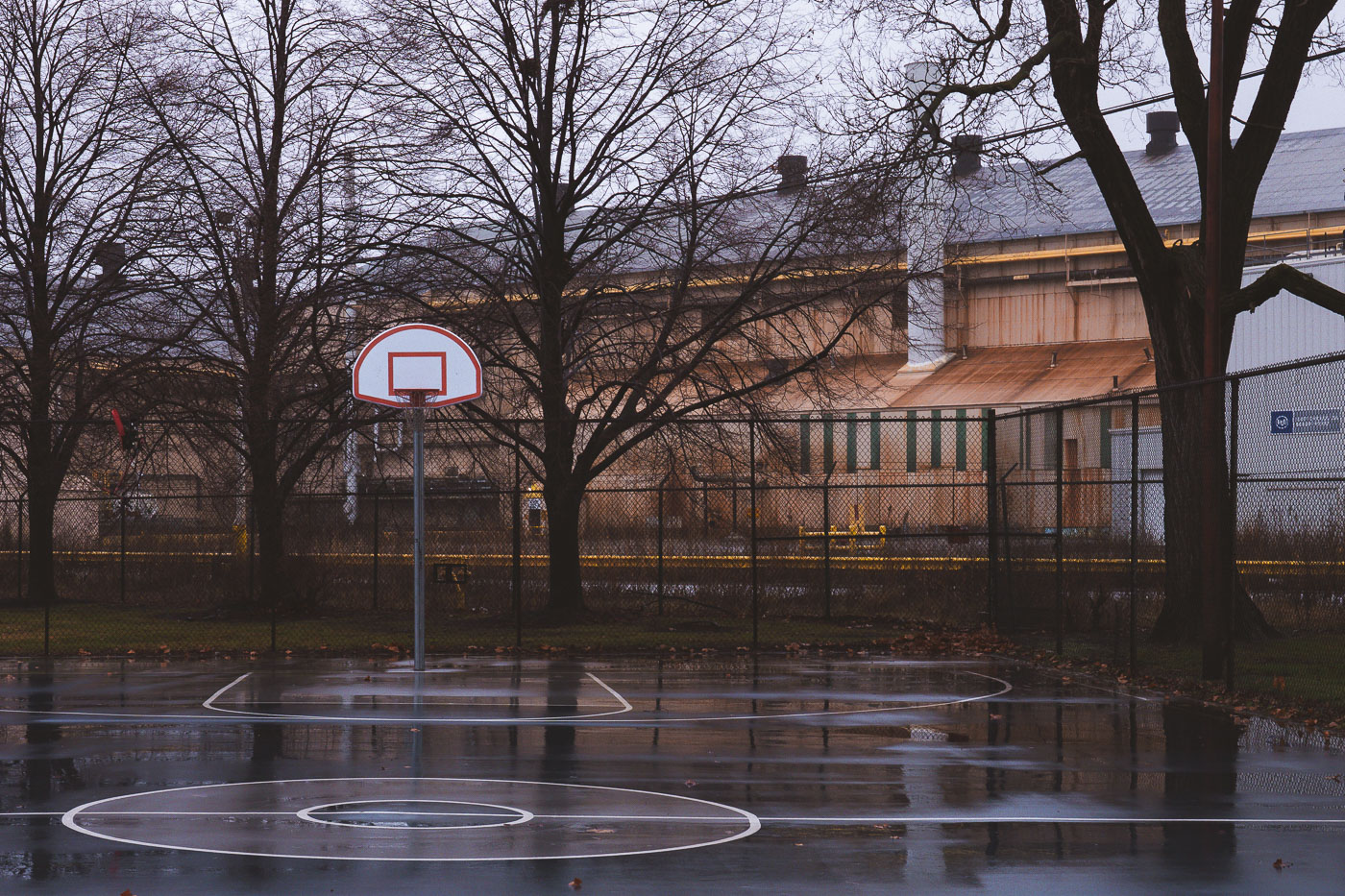 A wet basketball court with a hoop stands beside industrial buildings in Marktown, Indiana, a historic company town.