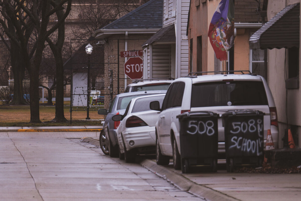 A stop sign on Spruce Ave in East Chicago, Indiana, with two trash bins marked "508 SCHOOL" at the curb.