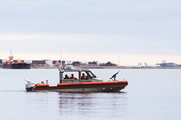 U.S. Coast Guard Patrol, Boston Harbor 4 A U.S. Coast Guard response boat moves steadily across Boston Harbor on a calm, overcast day, with Logan International Airport visible in the distance. The aluminum and orange-hulled vessel, equipped with mounted security equipment, is part of routine maritime patrol operations ensuring the safety of one of New England’s busiest ports. Behind it, commercial ships and aircraft highlight the harbor’s strategic role as a hub for both air and sea transport serving the Greater Boston area.
