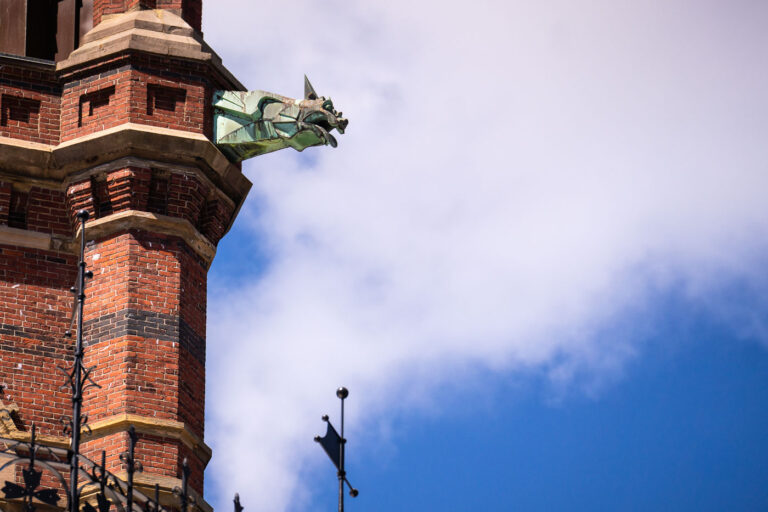 Gargoyle Detail at Harvard Memorial Hall 2 A copper gargoyle juts from the ornate brick tower of Harvard’s Memorial Hall in Cambridge, Massachusetts—a striking detail of the Gothic Revival architecture completed in the late 19th century. Designed by Ware & Van Brunt, the hall commemorates Harvard alumni who fought for the Union in the Civil War. The gargoyle’s oxidized green patina and sharp geometric form contrast against the red brick and open sky, blending medieval-inspired craftsmanship with the American collegiate Gothic style that defines much of Harvard’s architectural legacy.