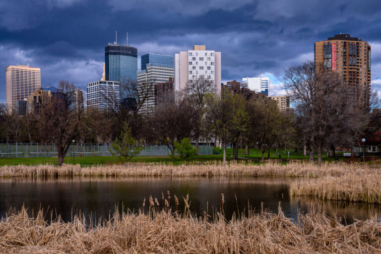 Minneapolis Skyline Over Loring Park Before a Storm 2 Dark storm clouds roll over downtown Minneapolis in this view from Loring Park, where the skyline’s mix of glass towers and historic brick buildings rises beyond the park’s quiet pond and reeds. The IDS Center, Capella Tower, and other high-rises dominate the horizon, their reflections muted in the pre-storm light. Taken during early spring, the trees remain bare while the surrounding grass begins to green, creating a striking contrast between nature’s seasonal transition and the geometric permanence of the city skyline.