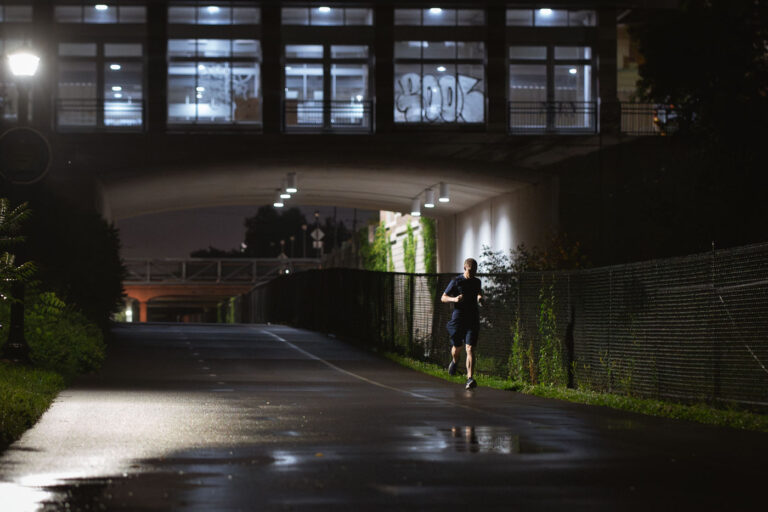 Jogger on Midtown Greenway, Minneapolis, After Rain 2 A lone jogger uses the Midtown Greenway in Minneapolis after a rain shower. This 5.7-mile paved trail follows the path of a former railroad line, converted into a public corridor in the late 1990s. The Greenway serves as a vital artery for non-motorized transportation and recreation, connecting diverse neighborhoods across South Minneapolis. The presence of graffiti on the overpass above suggests the ongoing cultural life of the urban environment.