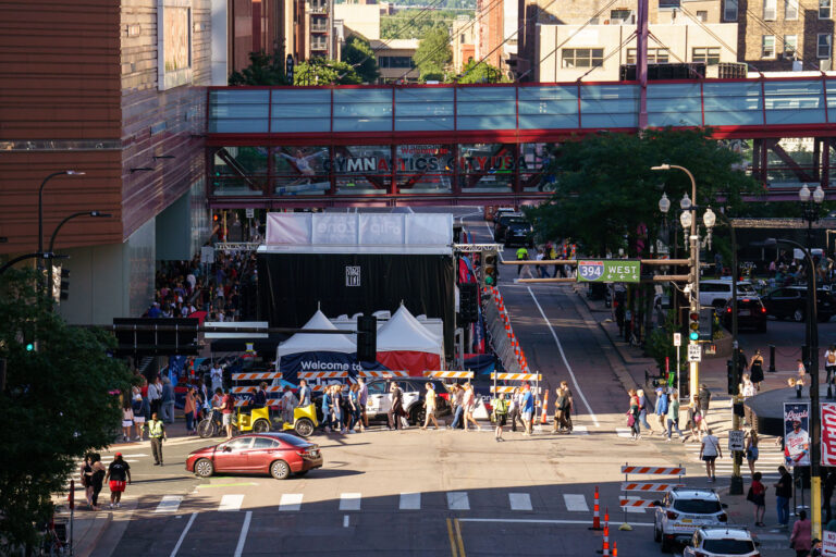US Gymnastics Olympic Trials at Target Center, Minneapolis 1 Pedestrians gather outside the Target Center in Minneapolis during the US Gymnastics Olympic Trials. A skybridge above the street displays "GYMNASTICS CITY USA" and images of gymnasts.