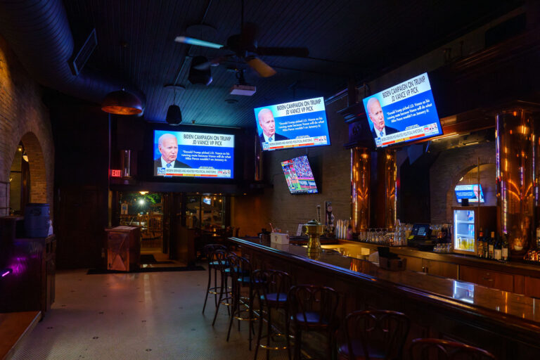 Televised Coverage of the 2024 Republican National Convention at 4 Televisions inside a Minneapolis bar show live coverage of the 2024 Republican National Convention, with news networks focusing on the Biden campaign’s response to Donald Trump’s selection of J.D. Vance as his running mate. The empty bar, dimly lit and lined with copper fixtures, reflects the late-night quiet after a long day of political events broadcast nationwide. The scene illustrates how political moments ripple far beyond the convention floor, reaching everyday public spaces where national politics become part of casual conversation.