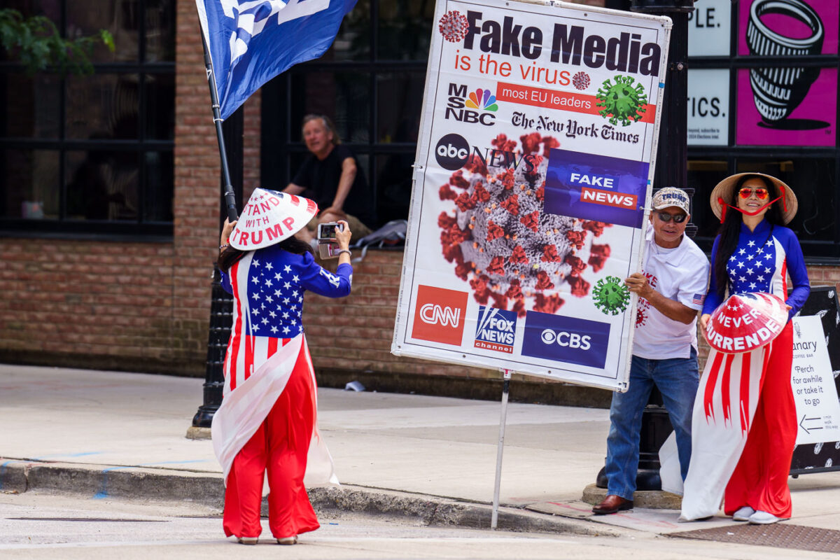 Man holds up a sign that reads "Fake media is the virus" and women hold hats that say "Stand with trump" "Never surrender". Never surrender began being used by supporters after Trump surrendered to Georgia authorities.