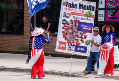 Man holds up a sign that reads "Fake media is the virus" and women hold hats that say "Stand with trump" "Never surrender". Never surrender began being used by supporters after Trump surrendered to Georgia authorities.