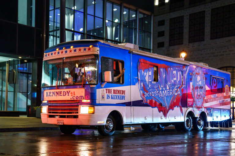 Kennedy 2024 Campaign Bus Parked Outside the RNC in Milwaukee 1 A brightly painted campaign bus promoting Robert F. Kennedy Jr.’s 2024 presidential bid sits outside a downtown Milwaukee venue during the Republican National Convention. The vehicle, wrapped in patriotic red, white, and blue artwork, bears slogans such as “The Remedy is Kennedy” and “Heal the Divide,” part of his campaign’s messaging focused on unity and reform.The bus’s retro RV design, complete with hand-painted lettering and American flag motifs, stands out against the modern glass architecture nearby. Its presence during the RNC reflected the independent candidate’s cross-party outreach and symbolic effort to engage voters across the political spectrum while touring key cities nationwide.