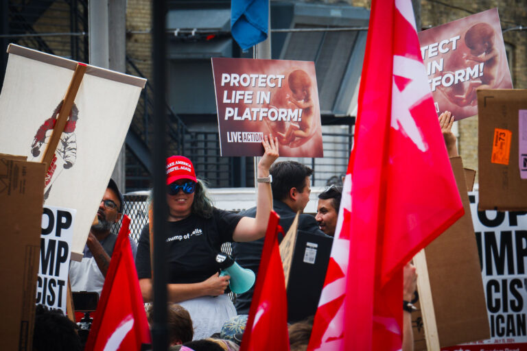 RNC Sign Protest Life In Platform 3 A woman wearing a "Make America Gospel Again" yells through a megaphone holding up a "Protest Life In Platform!" sing at the 2024 Republican National Convention.