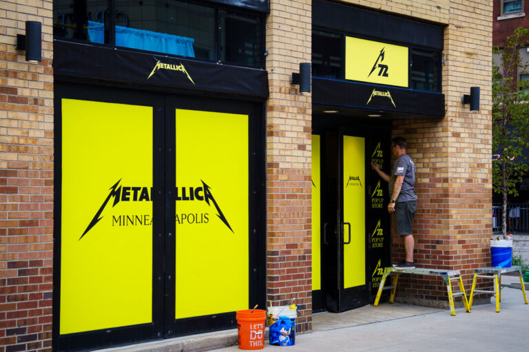 Metallica Pop-Up Store Installation, Minneapolis — August 2024 3 A worker installs bright yellow signage for a temporary Metallica pop-up store in downtown Minneapolis ahead of the band’s summer 2024 performances. The shop featured exclusive merchandise tied to Metallica’s 72 Seasons tour, one of the group’s largest North American runs in years. Pop-up locations like this were part of a coordinated campaign in major cities, connecting fans to the band’s tour stops with limited releases and local collaborations.