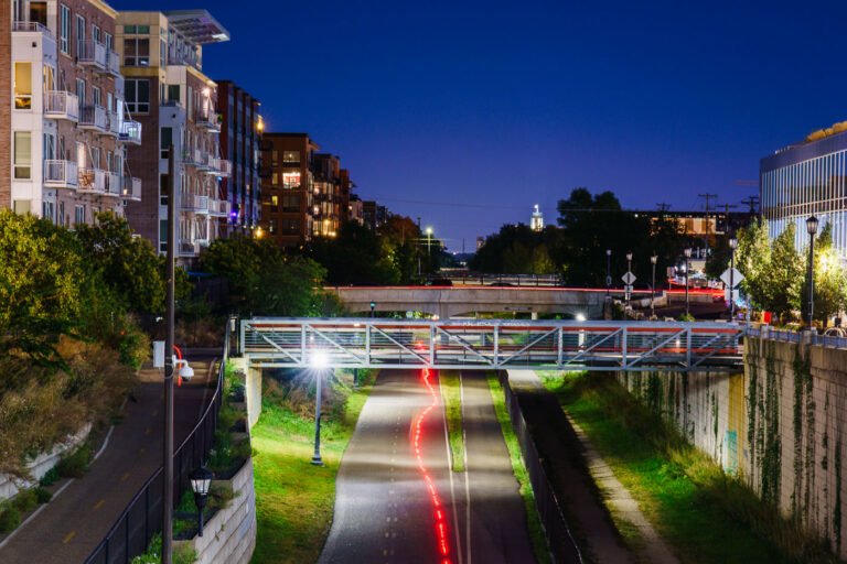 Midtown Greenway at Night – Minneapolis, MN 3 The Midtown Greenway runs beneath a series of bridges in south Minneapolis, shown here during evening hours with red tail lights from passing cyclists marking the trail’s path. Originally a railroad corridor built in the early 20th century, the Greenway was converted into a protected urban bike and pedestrian route in the early 2000s. Flanked by apartments, offices, and green spaces, it serves as a major east–west transportation corridor connecting Uptown to the Mississippi River while supporting Minneapolis’s commitment to sustainable transit.