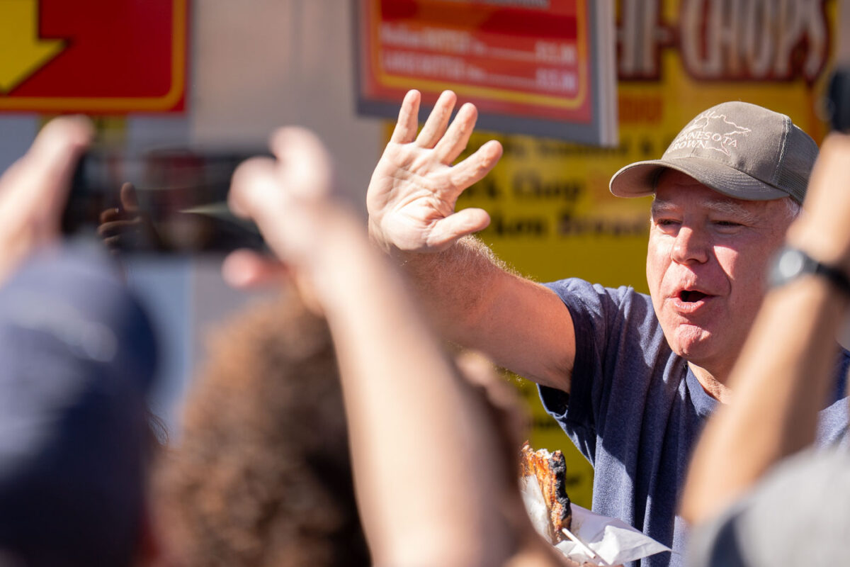 Tim Walz waves to those gathered outside the dairy building after biting into his Pork Chop On A Stick at the Minnesota State Fair on September 1, 2024.