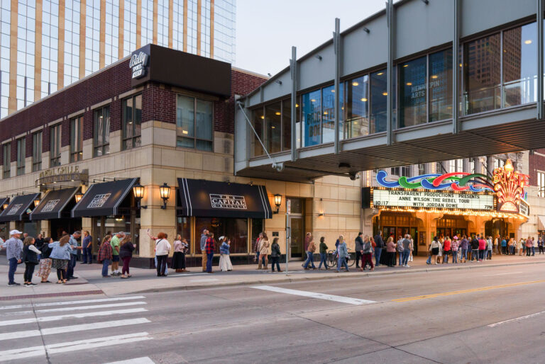 A line of concertgoers waits outside the historic State Theatre in downtown Minneapolis for a performance by Anne Wilson. The marquee indicates the event took place on October 17th, featuring "Anne Wilson: The Rebel Tour" with Jordan Rowe. The State Theatre, a landmark on Hennepin Avenue, has hosted numerous performances and events since its opening. Adjacent to the theater is The Capital Grille, a restaurant, and the building also houses Bally Sports North, indicating a mix of entertainment and commercial uses in this urban core.