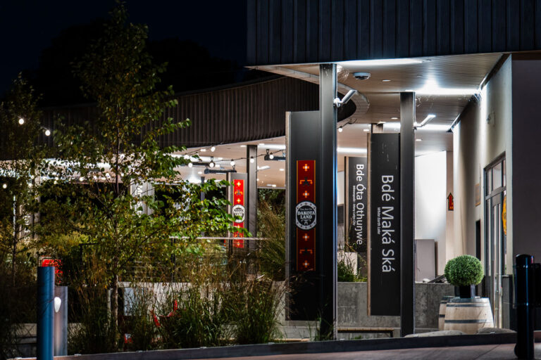 Bde Maka Ska Pavilion at Night — Minneapolis, Minnesota 4 The modern pavilion at Bde Maka Ska glows softly under evening light, its signage prominently honoring the lake’s Dakota name — “Bdé Óta Othúŋwe,” meaning “Many Village Place.” This site, located in southwest Minneapolis, holds deep historical and cultural significance as part of Dakota homeland, long before it was renamed “Lake Calhoun” in the 19th century. After years of advocacy by Indigenous groups and community leaders, the lake officially reclaimed its Dakota name in 2018, marking a shift toward recognizing Minnesota’s original peoples and languages. The illuminated pavilion stands today as both a gathering space and a statement of cultural restoration, blending modern urban design with respect for ancestral land and history.