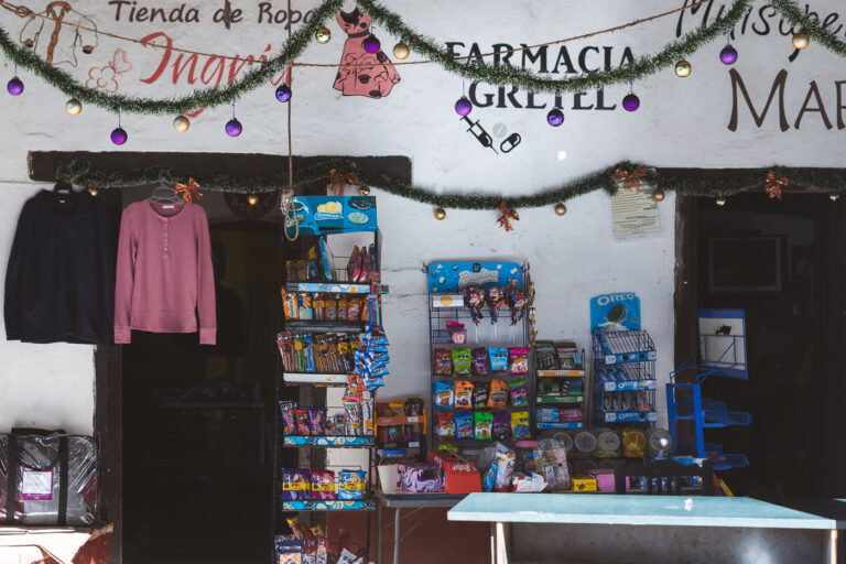 Farmacia Gretel, San Sebastián del Oeste 3 A small village pharmacy and convenience shop in San Sebastián del Oeste, Jalisco, displays everyday items from candy and cookies to basic clothing under hand-painted signs and Christmas garlands. The dual storefront—Farmacia Gretel and Tienda de Ropa Ingrith—reflects the multipurpose nature of businesses in this mountain town, where colonial-era buildings with thick adobe walls still house family-run enterprises. San Sebastián’s preserved architecture and slow rhythm have made it one of Mexico’s officially designated Pueblos Mágicos, drawing visitors seeking a glimpse of traditional Sierra Madre life.