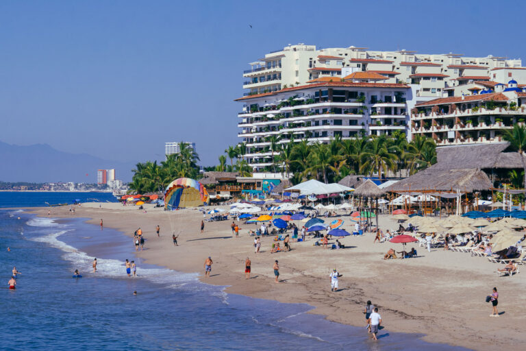 The densely layered hillsides of Puerto Vallarta rise above Playa de los Muertos, one of the city’s most iconic and active beachfronts. The shoreline is packed with rows of umbrellas and sunbathers, backed by mid-century and modern resort towers stacked against the green slopes of the Sierra Madre Occidental. Fishermen, swimmers, and jet-ski riders share the bay, while pelicans glide overhead. The neighborhood of Zona Romántica just inland mixes narrow cobblestone streets with lively bars and cafes, making this stretch of the Bahía de Banderas one of Mexico’s most photographed coastal skylines and a focal point of everyday life for both locals and visitors.