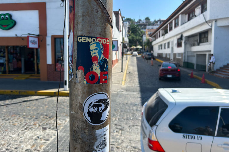 Political Stickers on a Street Pole, Puerto Vallarta 3 A pair of weathered stickers cling to a rusted street pole along a cobblestone street in Puerto Vallarta, Mexico. One features a political caricature with the word “GENOCIDE,” while the other, a minimalist face design, sits just below. Cars and pedestrians move through the narrow street lined with traditional whitewashed buildings, tiled roofs, and small local shops. The scene reflects how political expression and street art intermingle in everyday urban spaces, even in tourist-heavy coastal towns.