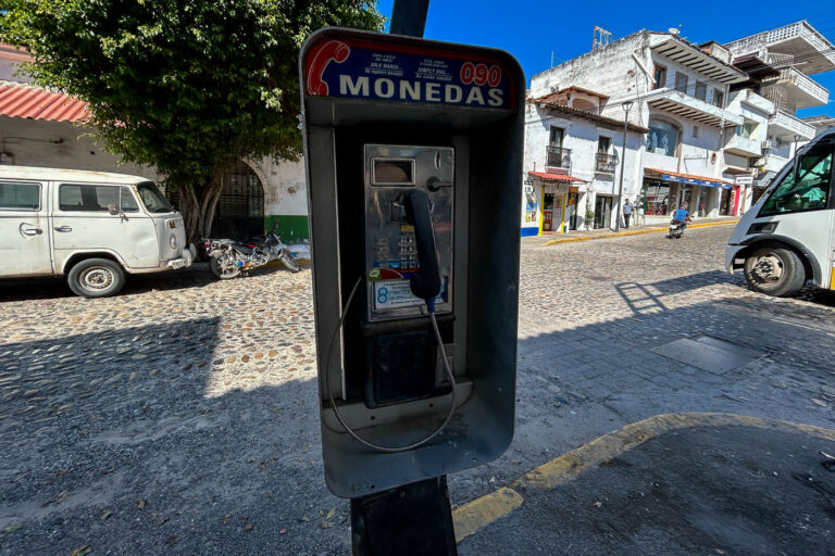 Coin Payphone on Calle Hidalgo, Puerto Vallarta 3 A coin-operated public payphone marked “Monedas 090” stands on a cobblestone street in central Puerto Vallarta, a reminder of the pre-digital era still visible in everyday urban life. Behind it, a mix of aging vehicles, small storefronts, and whitewashed buildings with red-tiled roofs reflect the city’s blend of local tradition and evolving modernity. These relics of analog communication, though seldom used today, remain scattered across Mexico’s towns and resort cities — quiet artifacts of an infrastructure that once connected travelers and locals alike.