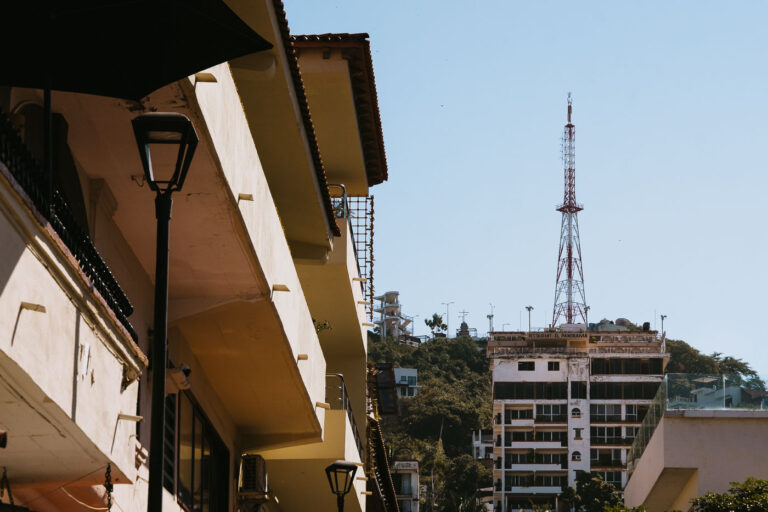 Hillside Towers of Puerto Vallarta 3 A narrow street in Puerto Vallarta’s Zona Romántica angles upward toward the city’s steep green hills, where a red-and-white radio tower crowns a weathered building labeled Restaurante El Panorama. The scene captures the city’s layered verticality—low stucco terraces with clay roofs in the foreground give way to midcentury apartment towers clinging to the slopes. The dense mix of balconies, street lamps, and satellite dishes reflects Puerto Vallarta’s blend of resort development and lived-in coastal community, where mountain terrain meets compact urban architecture.