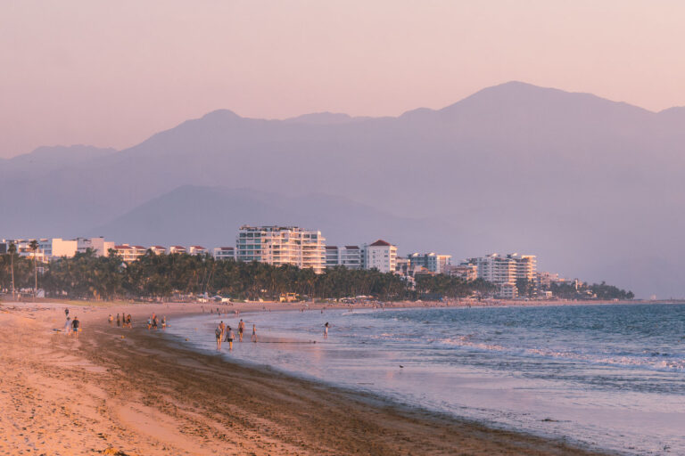 Coastal Walk at Nuevo Vallarta 3 Along the long shoreline of Nuevo Vallarta, visitors stroll the water’s edge where the resort towers of Riviera Nayarit meet the Pacific. The line of hotels and condominiums stretches toward Puerto Vallarta in the distance, marking one of Mexico’s most developed coastal corridors. Behind the beachfront skyline rise the forested slopes of the Sierra Madre Occidental, framing the region’s distinctive geography—where mountains descend sharply into the sea and urban leisure merges with natural backdrop.
