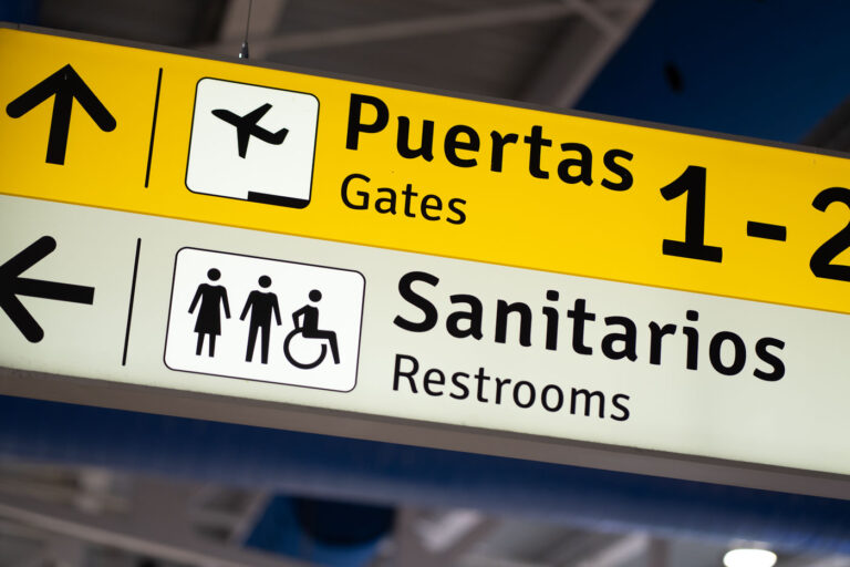 Bilingual Airport Signage in Puerto Vallarta Terminal 1 A close-up of bilingual directional signage inside Puerto Vallarta International Airport shows clear guidance for travelers in both Spanish and English. The yellow and white board directs passengers toward Gates 1–2 (“Puertas”) and nearby restrooms (“Sanitarios”), featuring internationally recognized icons for accessibility. This simple yet essential detail reflects Mexico’s effort to accommodate global visitors arriving through one of the country’s busiest coastal gateways.