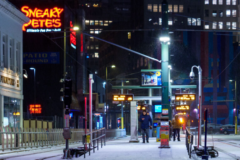 Winter Night at Warehouse District Station — Minneapolis, Janu 4 Under the glow of neon and transit lights, a few bundled-up pedestrians wait for the train at Warehouse District/Hennepin Avenue Station in downtown Minneapolis on a frigid January night. The temperature hovered in the single digits as light snow dusted the platform and tracks. The glowing red sign for Sneaky Pete’s — a longtime nightlife landmark just off 1st Avenue — adds a warm contrast to the blue-gray tones of the winter scene. The photo captures the resilient quiet of Minneapolis transit life in deep winter, where late-night riders and flickering lights define the city’s pulse long after rush hour ends.