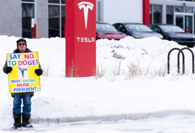 A man stands with a sign reading "Say No To Doge! Who Elected Musk President?" outside a Tesla facility in Minneapolis.