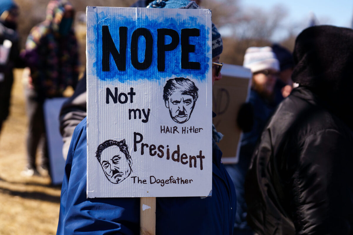 Protester holding up a sign that reads 

"Nope Not my President"

"Hair Hitler"
"The Dogefather"

Protesters have gathered each Saturday for weeks now at this Tesla Facility just outside of Minneapolis (Golden Valley).