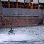 A man on a jet ski in the port of Miami text to large container ships.