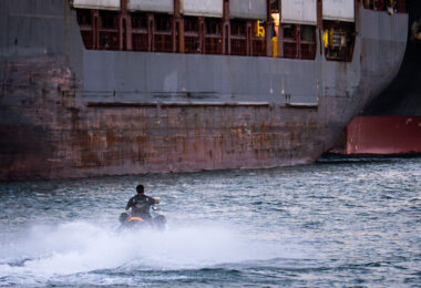 A man on a jet ski in the port of Miami text to large container ships.
