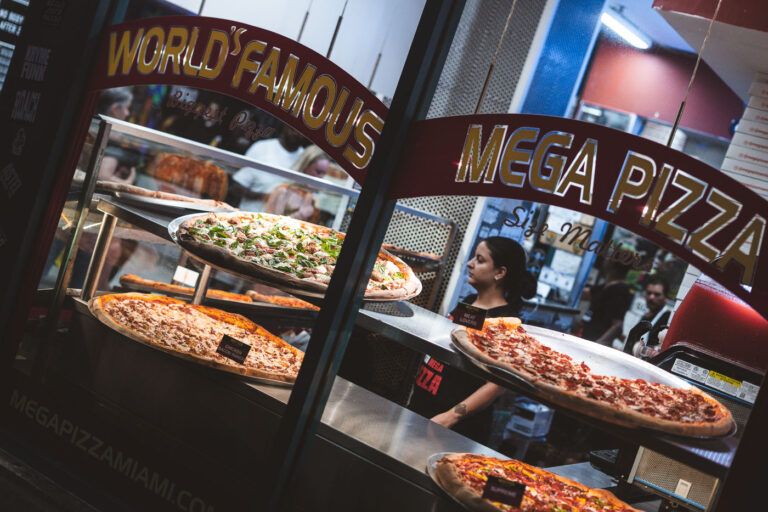 Late-Night Pizza at Mega Pizza, Miami 4 A worker at Mega Pizza serves giant slices through the restaurant’s glass storefront in Miami, where enormous pizzas are proudly displayed under the “World Famous” and “Size Matters” signage. The stacked trays of oversized slices—ranging from chicken bacon ranch to meat lovers—showcase the shop’s signature flair for indulgence. The neon-lit interior and street reflections outside capture the casual energy of Miami’s late-night dining scene.