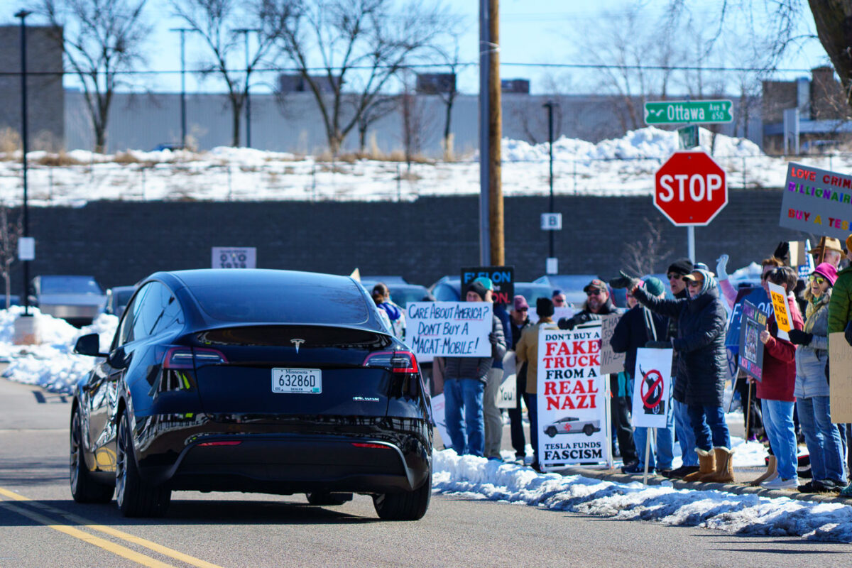 A Tesla car drives by a group of protesters in Golden Valley (Minneapolis). The protesters have been gathering for weeks as part of "Tesla Takedown" protests.