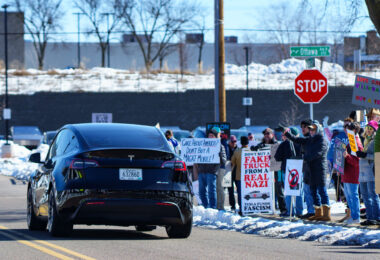 A Tesla car drives by a group of protesters in Golden Valley (Minneapolis). The protesters have been gathering for weeks as part of "Tesla Takedown" protests.