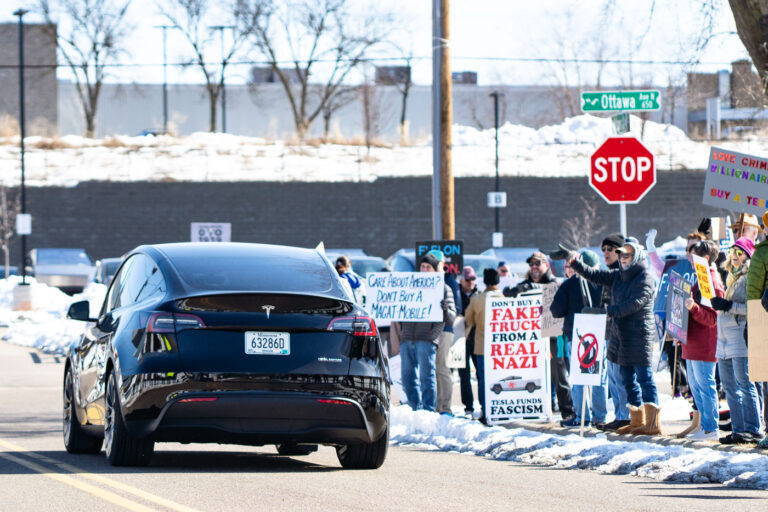 Tesla car in front of protesters in Minneapolis 2 A Tesla car drives by a group of protesters in Golden Valley (Minneapolis). The protesters have been gathering for weeks as part of "Tesla Takedown" protests.