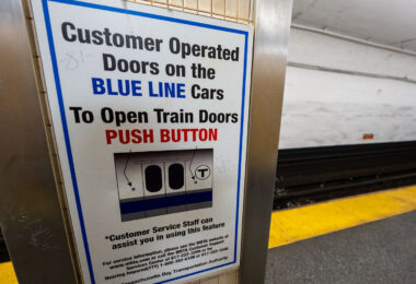 A platform sign explaining customer-operated doors on Blue Line trains of the Massachusetts Bay Transportation Authority. Unlike many rapid-transit systems where doors open automatically at every stop, portions of the MBTA Blue Line fleet require passengers to press a button to open the doors once the train is stopped. This practice dates back to older car designs and cold-weather operating considerations, helping retain heat in winter and reduce unnecessary door cycling. The sign reflects the MBTA’s continued reliance on legacy rolling stock and the need for clear, standardized instructions in stations with high passenger turnover.