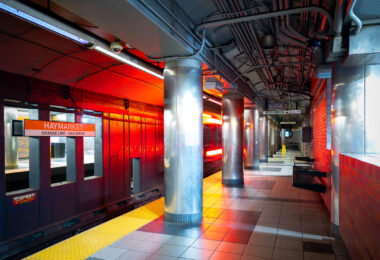 Haymarket Station in downtown Boston, one of the oldest transit locations in the city, serving riders since the late 19th century in various forms. The current underground station opened in 1971 as part of the MBTA’s modernization of the Orange Line, replacing earlier elevated structures that once ran through the area. The station sits at the edge of the historic Haymarket district, long associated with public markets and transportation links between downtown and Boston’s North End. The cylindrical steel columns and low-ceilinged platforms reflect mid-20th-century transit design, while the signage and lighting document decades of incremental updates to keep the station functional within one of the system’s busiest transfer points.
